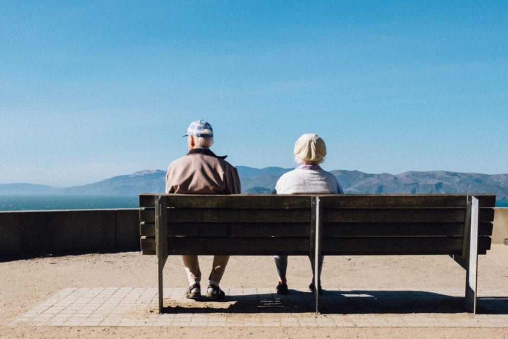 et ældre pensionist par sidder på en bænk og kigger på en blå himmel og bjerge i baggrunden